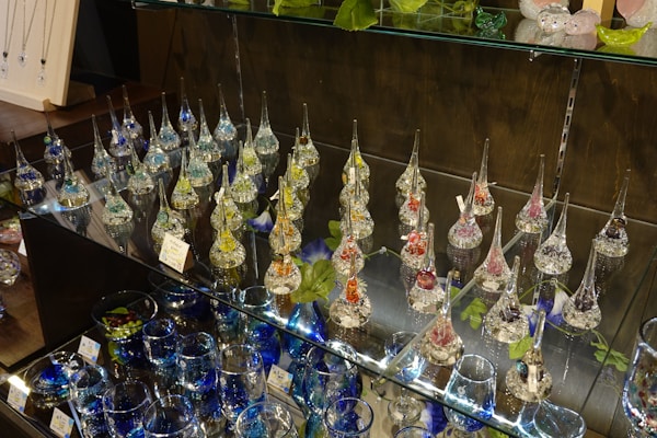 Variety of delicate glass ornaments with long, slender necks displayed on glass shelves. The ornaments are filled with colorful materials like beads or glitter, creating a shimmering effect under the light. Several leaves and other decorative glass items such as bowls and marbles are also arranged on the shelves.