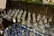 Variety of delicate glass ornaments with long, slender necks displayed on glass shelves. The ornaments are filled with colorful materials like beads or glitter, creating a shimmering effect under the light. Several leaves and other decorative glass items such as bowls and marbles are also arranged on the shelves.