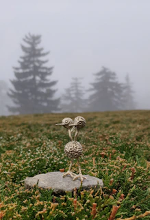 A small metal sculpture of two abstract bird-like figures stands on a flat rock, surrounded by dense, green foliage. In the background, several tall, blurry pine trees emerge through a thick layer of fog creating a mysterious and calm atmosphere.
