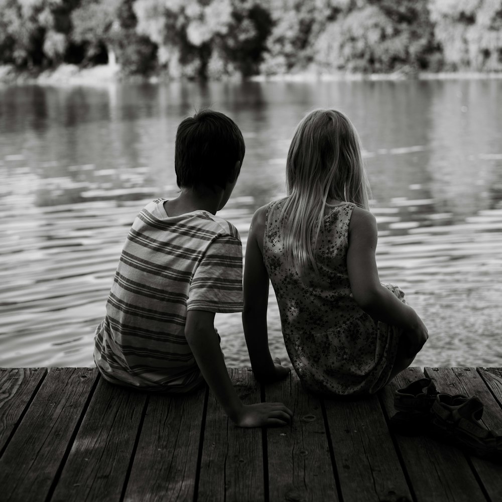 a couple of kids sitting on a dock next to a body of water