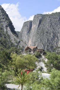 A rustic cabin is nestled among lush greenery, positioned high on a rocky hill. Towering cliffs dominate the background, creating a dramatic natural setting. The sky is mostly clear with a few fluffy clouds, adding to the serene and picturesque atmosphere. A red object and a white structure, possibly vehicles or storage units, are partially visible in the foreground surrounded by various types of vegetation.