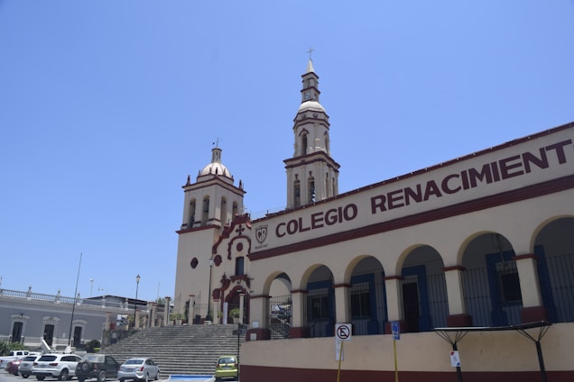 A building with two ornate towers and arched windows is seen under a clear blue sky. The building has the words 'Colegio Renacimiento' prominently displayed on its facade. Several parked cars are visible in the foreground.