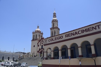 A building with two ornate towers and arched windows is seen under a clear blue sky. The building has the words 'Colegio Renacimiento' prominently displayed on its facade. Several parked cars are visible in the foreground.