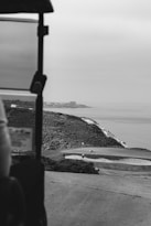 A serene golf course overlooking the ocean is visible, with three people on the green. A cliffside with lush vegetation extends toward the horizon, and a cityscape can be seen in the distance. The foreground includes part of a golf cart frame.