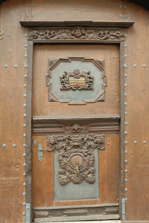 An ornately carved wooden door with intricate details, including floral patterns and cherub faces. The door features a plaque with an inscription, likely indicating a historical or commemorative message. The design reflects classical and elaborate craftsmanship.