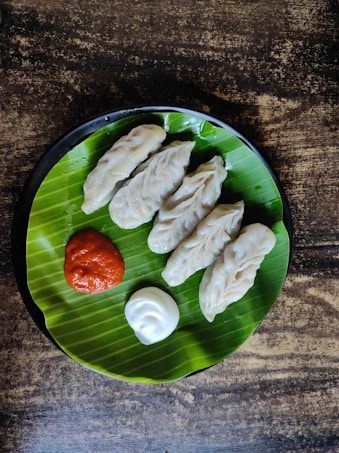 A black plate contains five dumplings neatly arranged on a vibrant green banana leaf, accompanied by a dollop of red sauce and a dollop of white sauce. The setup is placed on a rustic wooden surface, providing a contrast to the bright leaf and the subtle tones of the dumplings.