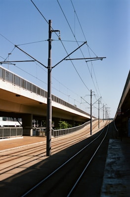 Railway tracks run under a clear blue sky alongside a light rail or tram system. The tracks are flanked by overhead electrical lines supported by poles. A concrete overpass stretches parallel to the tracks on the left side, with safety railings lining its path. There is a platform on the right, possibly for boarding, and sparse greenery visible in the distance.