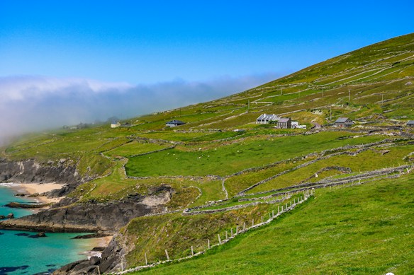 A scenic coastal landscape with vibrant green hillsides descending towards a rocky shore and azure sea. Stone fences divide the grassy areas, and several small houses dot the landscape. A misty sky adds a sense of depth and atmosphere to the scene.