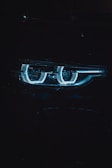 Close-up shot of a car headlight with a futuristic and sleek design, showcasing a distinctive blue LED ring pattern. The image is dimly lit, emphasizing the glow of the lights against the dark background.
