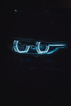 Close-up shot of a car headlight with a futuristic and sleek design, showcasing a distinctive blue LED ring pattern. The image is dimly lit, emphasizing the glow of the lights against the dark background.