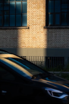 A brick wall with large blue-tinted windows in an industrial-looking building. The sunlight casts a sharp shadow across the wall, creating a contrast between light and shadow. In the foreground, a car with reflective surfaces adds dynamic lines and reflections.