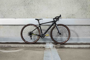 A modern road bicycle with a sleek black frame and thin tires is positioned against a textured, industrial concrete wall. The bike features drop handlebars and subtle accents, with yellow text and branding near the chain area. The setting suggests an urban environment with muted, understated tones.