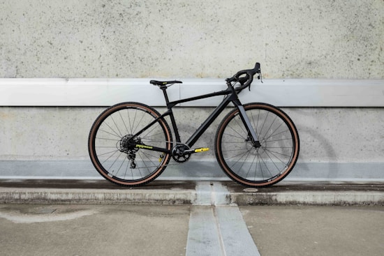 A modern road bicycle with a sleek black frame and thin tires is positioned against a textured, industrial concrete wall. The bike features drop handlebars and subtle accents, with yellow text and branding near the chain area. The setting suggests an urban environment with muted, understated tones.