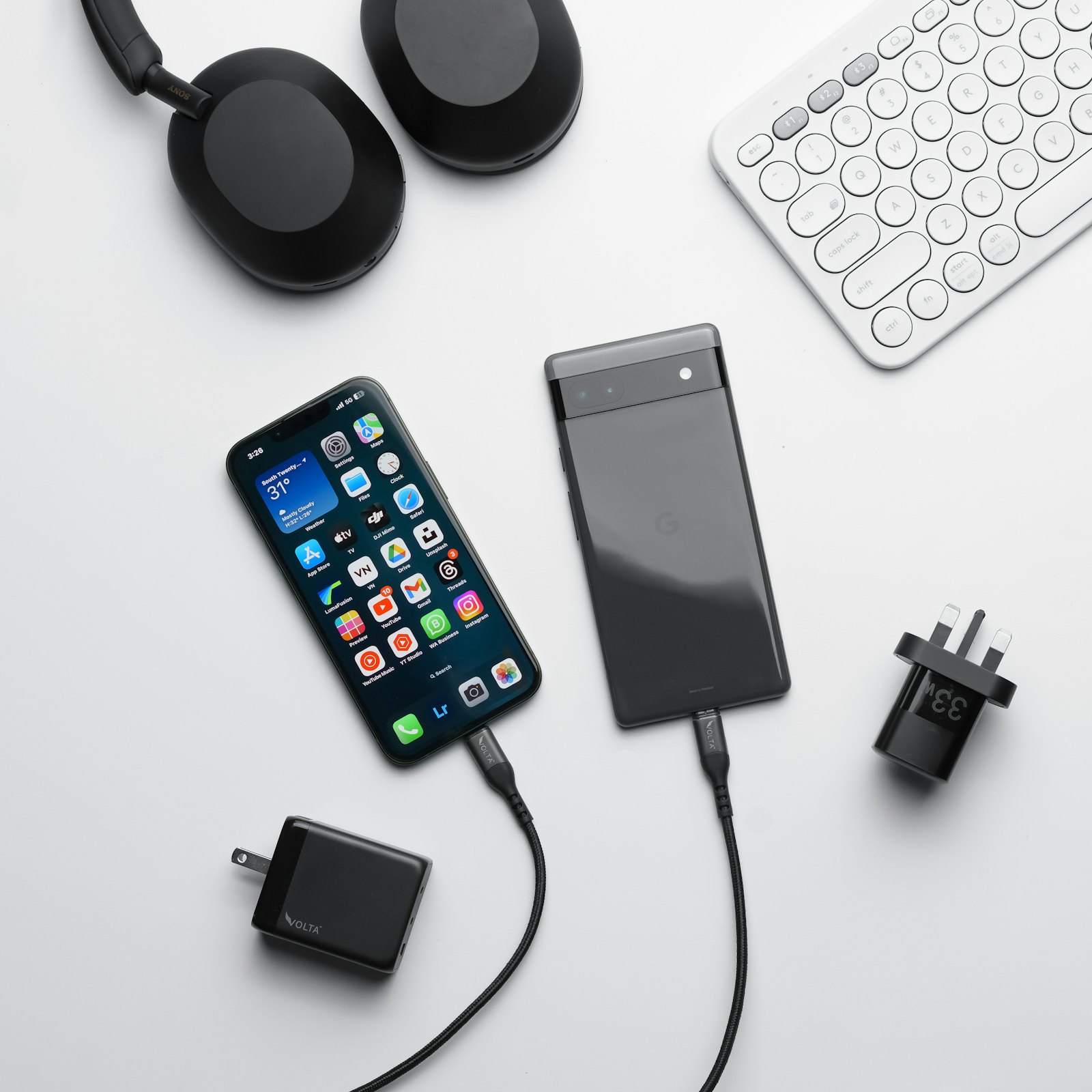 A phone, wireless headphones, a keyboard and chargers laid out on a white desk