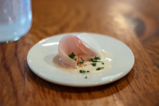 A small, elegantly presented dish sits on a white plate. The dish consists of thinly sliced, translucent fish, garnished with finely chopped herbs and a creamy white sauce. It is set on a wooden table with a subtle reflection visible on the surface.