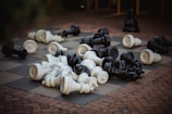 Large chess pieces are scattered across an outdoor chessboard, with both black and white pieces lying on their sides. The board is set on a brick patio, creating a contrast between the checkered pattern of the board and the texture of the bricks.