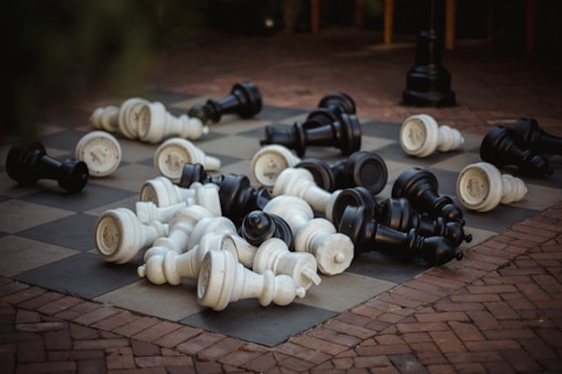 Large chess pieces are scattered across an outdoor chessboard, with both black and white pieces lying on their sides. The board is set on a brick patio, creating a contrast between the checkered pattern of the board and the texture of the bricks.