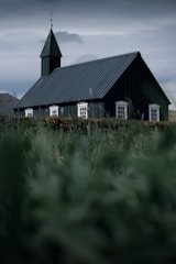 A dark, black wooden church stands with a tall steeple and small white-framed windows against a backdrop of cloudy skies. Lush green grass fills the foreground, leading up to the church structure, which exudes a rustic and historic charm. The overall atmosphere is serene and slightly moody due to the overcast weather.