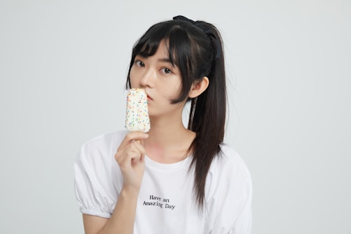 A young person with a ponytail is holding a popsicle with colorful sprinkles. They are wearing a white t-shirt with the text 'Have an Amazing Day' printed on it. The background is plain and light-colored.