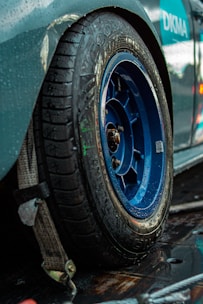 A close-up view of a car tire with a blue alloy wheel, showing clear water droplets on the surface. The tire is on a slightly reflective, wet surface, suggesting recent rain or washing. A secured strap is visible near the tire, indicating the car might be fastened to a trailer or lift. There are parts of the car body visible, primarily in green and blue shades.