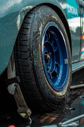 A close-up view of a car tire with a blue alloy wheel, showing clear water droplets on the surface. The tire is on a slightly reflective, wet surface, suggesting recent rain or washing. A secured strap is visible near the tire, indicating the car might be fastened to a trailer or lift. There are parts of the car body visible, primarily in green and blue shades.