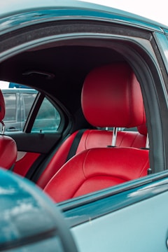 The interior of a car features vibrant red leather seats against a backdrop of a neutral-toned car exterior. The door frame and window are visible, with a glimpse of a vehicle parked beside it.