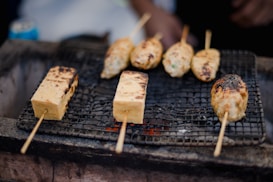 Skewered food items are being grilled on a mesh over an open flame. The skewers include rectangular tofu blocks and oblong meat or vegetable patties, each showing signs of charring from the grilling process.