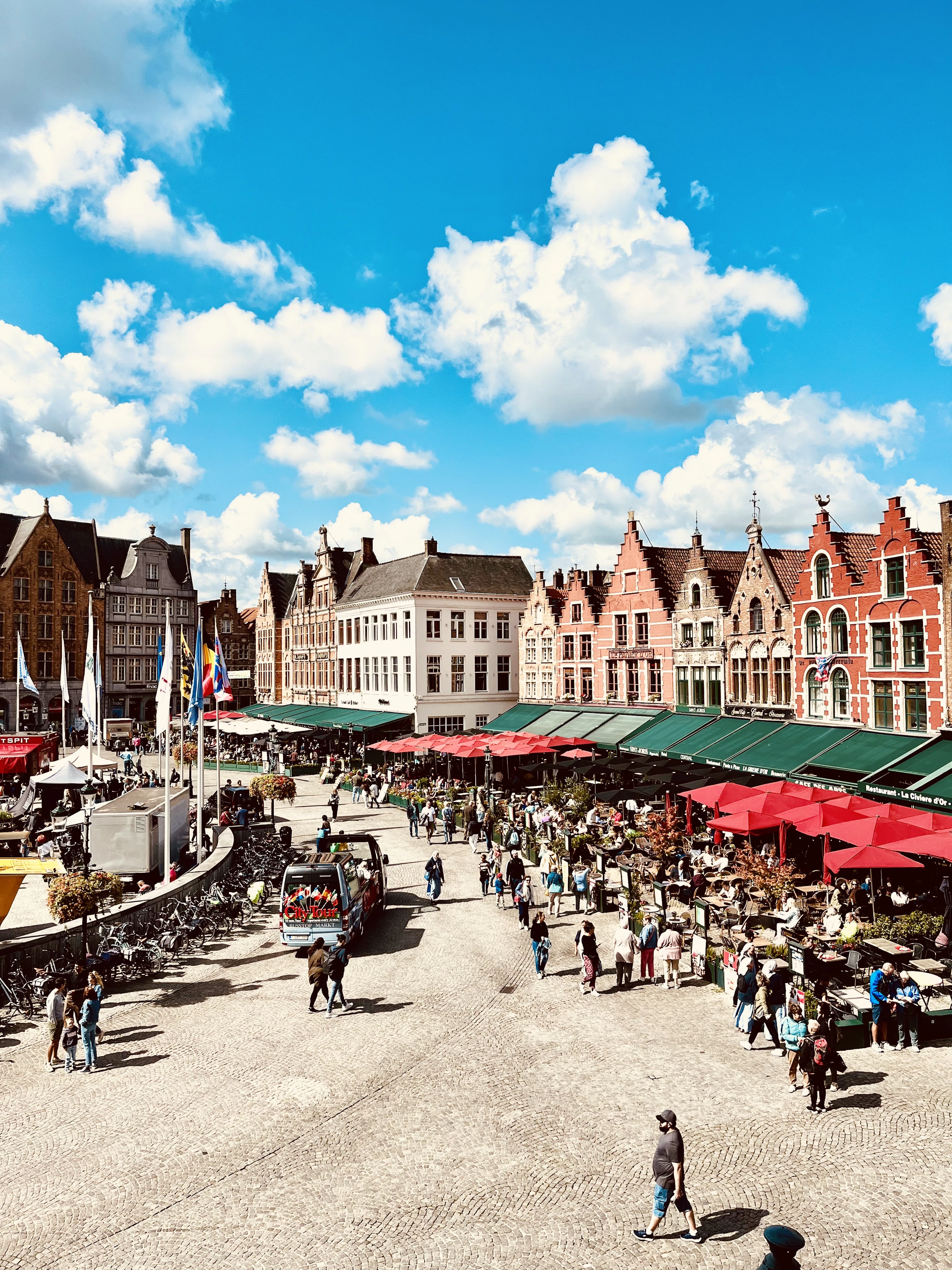 Market stalls under red awnings fill Bruges' Markt square beneath colorful step-gabled houses and a bright sky.