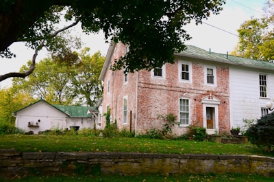 A rustic brick house with an adjoining white extension sits in a lush green yard surrounded by mature trees. The roof is green, and there is a small outbuilding with a matching green roof nearby. The foreground features a stone wall and a well-maintained lawn.