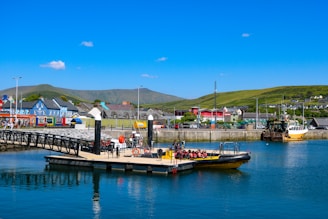 A vibrant harbor scene featuring a small dock with a yellow boat moored at the waterfront. Several people are seated on the boat, wearing life vests. In the background, colorful buildings line the waterfront, and green hills stretch into the distance under a clear blue sky.