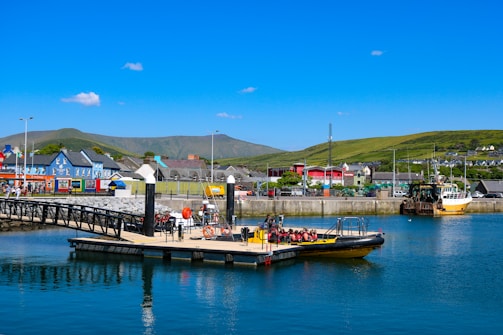 A vibrant harbor scene featuring a small dock with a yellow boat moored at the waterfront. Several people are seated on the boat, wearing life vests. In the background, colorful buildings line the waterfront, and green hills stretch into the distance under a clear blue sky.