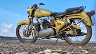 A classic Royal Enfield motorcycle with a vintage design in mustard color is parked on a ground with gravel and shells visible in the foreground. The motorcycle has a prominent '350' written on it and the background shows a clear blue sky with some clouds and distant foliage.