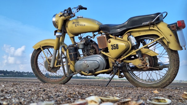 A classic Royal Enfield motorcycle with a vintage design in mustard color is parked on a ground with gravel and shells visible in the foreground. The motorcycle has a prominent '350' written on it and the background shows a clear blue sky with some clouds and distant foliage.