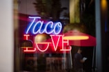A neon sign displaying the words 'Taco Love' in bright blue and red colors is prominently featured. In the background, blurred images of a person under a red umbrella and what appears to be historical posters add depth to the scene.