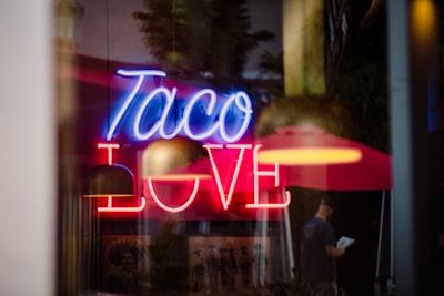 A neon sign displaying the words 'Taco Love' in bright blue and red colors is prominently featured. In the background, blurred images of a person under a red umbrella and what appears to be historical posters add depth to the scene.