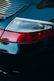 A close-up view of a car's rear end, focusing on the tail light and rear panel with shiny black paint reflecting light. The tail light features bright red and white sections, and the sleek design suggests a high-end model. The glossy surface captures reflections and gives a sense of luxury and sophistication.
