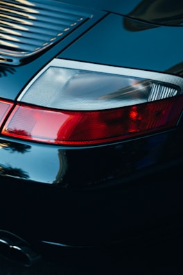 A close-up view of a car's rear end, focusing on the tail light and rear panel with shiny black paint reflecting light. The tail light features bright red and white sections, and the sleek design suggests a high-end model. The glossy surface captures reflections and gives a sense of luxury and sophistication.