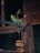 A green and yellow parakeet perches on a branch inside a wooden aviary. The bird exhibits intricate feather patterns. A second parakeet is visible in the background. A wooden toy or decoration hangs next to the branch.