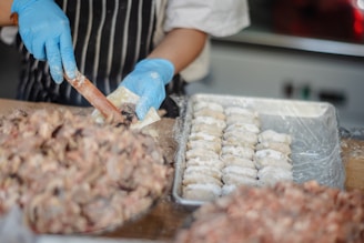 A person wearing blue gloves and a striped apron is preparing food, likely dumplings or pastries. They are handling dough and filling with a utensil. In the foreground, there is a large pile of mixture and a tray of neatly arranged, uncooked dumplings dusted with flour.