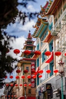 A vibrant street scene in a Chinatown, featuring traditional Chinese architectural elements such as colorful roofs and ornamental details. Numerous red lanterns are hanging across the street, adding a festive and cultural atmosphere. The buildings have intricate, multi-tiered roofs with red, green, and gold accents.