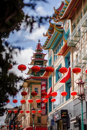 A vibrant street scene in a Chinatown, featuring traditional Chinese architectural elements such as colorful roofs and ornamental details. Numerous red lanterns are hanging across the street, adding a festive and cultural atmosphere. The buildings have intricate, multi-tiered roofs with red, green, and gold accents.