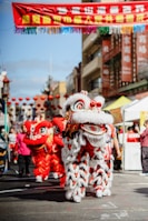 A vibrant street scene features a traditional lion dance performance, with two colorful lion costumes predominantly in red and white. The background shows urban buildings with Chinese banners, festive decorations, and lanterns lining the street.