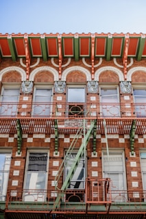 A vintage brick building facade featuring decorative arches and ornate metalwork. The structure includes a fire escape painted in a contrasting green, complementing the red and brown tones of the brickwork. The architectural details include sculpted elements above the windows and a grid pattern created by lighter bricks.
