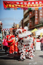 A vibrant street scene featuring a traditional Chinese lion dance performance with performers in bright red and white costumes, extravagant lion heads, and detailed decorations. Spectators can be seen on the sides, and red lanterns are strung across the street, adding to the festive atmosphere.