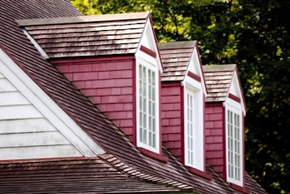 A series of three dormer windows protrude from a sloped roof clad in red shingles. Each dormer is painted in a deep red color with white window frames and trim. The main roof is covered in dark brown shingles, and there are green trees in the background, suggesting a lush, outdoor setting.