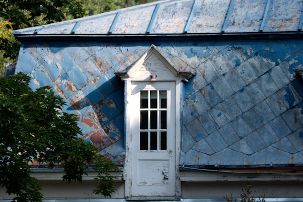 A weathered and old blue metal roof with visible patches of rust and peeling paint is highlighted by sunlight. Below, a small white-framed window with multiple panes is set into the roof structure. Tree branches with green leaves can be seen in the foreground, adding a touch of nature to the scene.