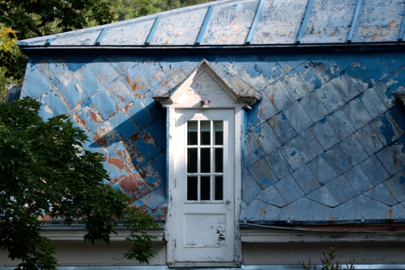 A weathered and old blue metal roof with visible patches of rust and peeling paint is highlighted by sunlight. Below, a small white-framed window with multiple panes is set into the roof structure. Tree branches with green leaves can be seen in the foreground, adding a touch of nature to the scene.