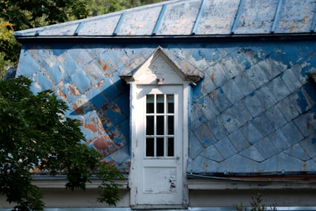 A weathered and old blue metal roof with visible patches of rust and peeling paint is highlighted by sunlight. Below, a small white-framed window with multiple panes is set into the roof structure. Tree branches with green leaves can be seen in the foreground, adding a touch of nature to the scene.