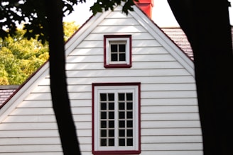 A white clapboard house with maroon trim and a steeply pitched roof. Two windows, one larger with multiple panes and one smaller above it, are framed by the contrasting trim. Trees partially frame the house, with some leaves casting shadows.