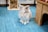 A fluffy white rabbit with long ears sits on a textured blue rug. To the right, there is a wooden structure resembling a small hutch or shelter. In the background, there's a vertical holder filled with hay.